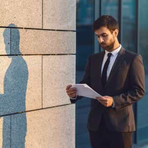 Serious young businessman in a classic suit reading advantageous business document in paper